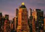 A cityscape at dusk shows the illuminated skyline of Manhattan. At the center stands One Worldwide Plaza, a tall building with a pyramid-shaped green roof topped by a glowing triangular light. The tower is surrounded by other high-rise buildings, their windows lit against the purple and orange evening sky.