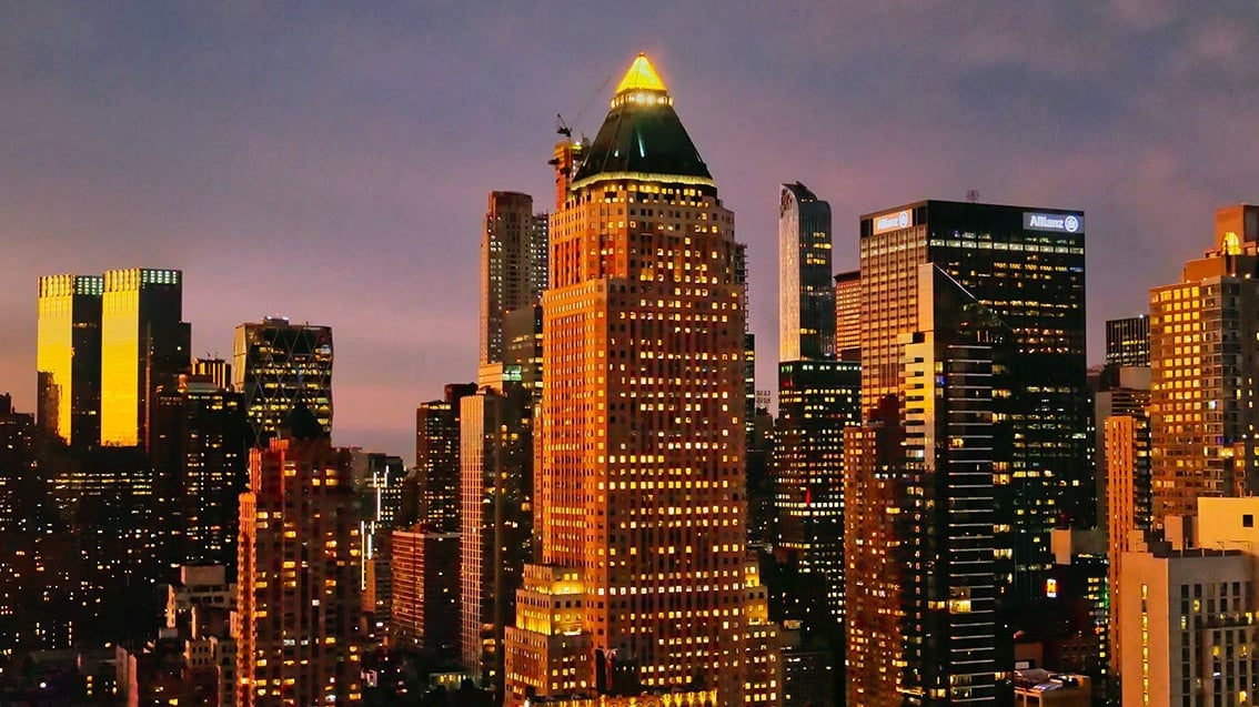 A cityscape at dusk shows the illuminated skyline of Manhattan. At the center stands One Worldwide Plaza, a tall building with a pyramid-shaped green roof topped by a glowing triangular light. The tower is surrounded by other high-rise buildings, their windows lit against the purple and orange evening sky.
