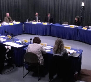 Members of the Arkansas PBS Commission at their quarterly meeting on June 5, seated at tables covered in blue cloth and arranged in a U shape with microphones, nameplates and papers in front of them in a studio with gray curtains.