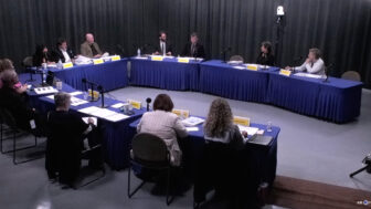 Members of the Arkansas PBS Commission at their quarterly meeting on June 5, seated at tables covered in blue cloth and arranged in a U shape with microphones, nameplates and papers in front of them in a studio with gray curtains.