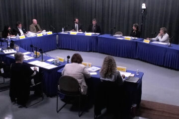 Members of the Arkansas PBS Commission at their quarterly meeting on June 5, seated at tables covered in blue cloth and arranged in a U shape with microphones, nameplates and papers in front of them in a studio with gray curtains.