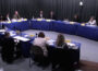 Members of the Arkansas PBS Commission at their quarterly meeting on June 5, seated at tables covered in blue cloth and arranged in a U shape with microphones, nameplates and papers in front of them in a studio with gray curtains.