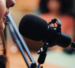 Close-up of a person speaking into a studio microphone, with blurred background suggesting a recording setting.