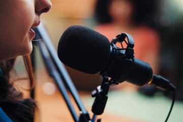 Close-up of a person speaking into a studio microphone, with blurred background suggesting a recording setting.