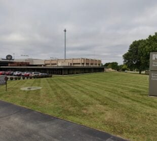 Exterior view of the Kentucky Educational Television headquarters, a low modern building with tan panels and large windows, a tall transmission tower rising behind it. A grassy lawn and parking lot with cars sit in the foreground, with a sign reading “KET The Kentucky Network, O. Leonard Press Telecommunications Center” near the sidewalk.