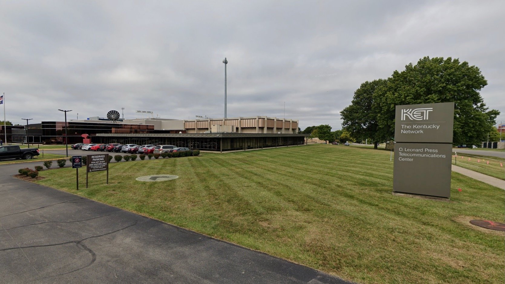 Exterior view of the Kentucky Educational Television headquarters, a low modern building with tan panels and large windows, a tall transmission tower rising behind it. A grassy lawn and parking lot with cars sit in the foreground, with a sign reading “KET The Kentucky Network, O. Leonard Press Telecommunications Center” near the sidewalk.