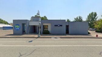Street view of KIXE PBS headquarters in Redding, California, a low gray building with the PBS Kids logo on the left and the KIXE PBS logo on the right, with a sidewalk and a streetlamp in front.