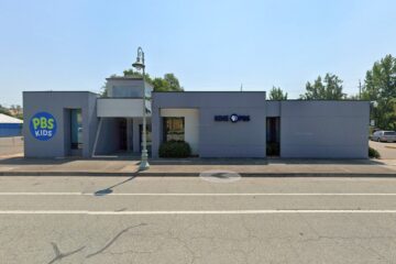 Street view of KIXE PBS headquarters in Redding, California, a low gray building with the PBS Kids logo on the left and the KIXE PBS logo on the right, with a sidewalk and a streetlamp in front.