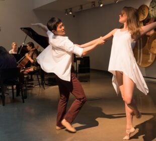 Two dancers, Ashley Thursby-Kern and Ryo Suzuki, perform in front of string musicians Annie Daigle, Rachel Grimes, Cecilia Huerta-Lauf, and Carrie RavenStem during a New Lens concert at the 21c Museum Hotel in Louisville, with woven basket art displayed on the wall behind them.