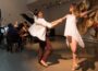 Two dancers, Ashley Thursby-Kern and Ryo Suzuki, perform in front of string musicians Annie Daigle, Rachel Grimes, Cecilia Huerta-Lauf, and Carrie RavenStem during a New Lens concert at the 21c Museum Hotel in Louisville, with woven basket art displayed on the wall behind them.