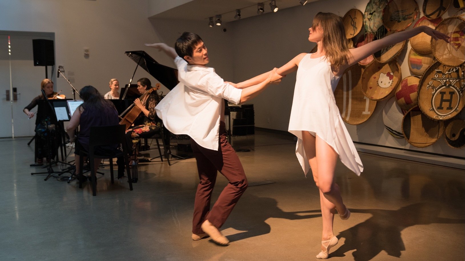Two dancers, Ashley Thursby-Kern and Ryo Suzuki, perform in front of string musicians Annie Daigle, Rachel Grimes, Cecilia Huerta-Lauf, and Carrie RavenStem during a New Lens concert at the 21c Museum Hotel in Louisville, with woven basket art displayed on the wall behind them.