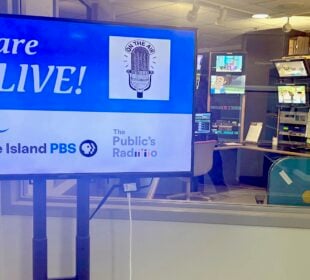 A television monitor in a control room displays the message “We are LIVE!” with a vintage microphone graphic and the words “On the Air.” Logos for Rhode Island PBS and The Public’s Radio appear below. Behind the monitor, through a window, television monitors and broadcasting equipment fill the control room.