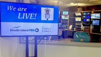 A television monitor in a control room displays the message “We are LIVE!” with a vintage microphone graphic and the words “On the Air.” Logos for Rhode Island PBS and The Public’s Radio appear below. Behind the monitor, through a window, television monitors and broadcasting equipment fill the control room.