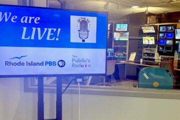 A television monitor in a control room displays the message “We are LIVE!” with a vintage microphone graphic and the words “On the Air.” Logos for Rhode Island PBS and The Public’s Radio appear below. Behind the monitor, through a window, television monitors and broadcasting equipment fill the control room.