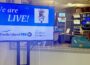 A television monitor in a control room displays the message “We are LIVE!” with a vintage microphone graphic and the words “On the Air.” Logos for Rhode Island PBS and The Public’s Radio appear below. Behind the monitor, through a window, television monitors and broadcasting equipment fill the control room.