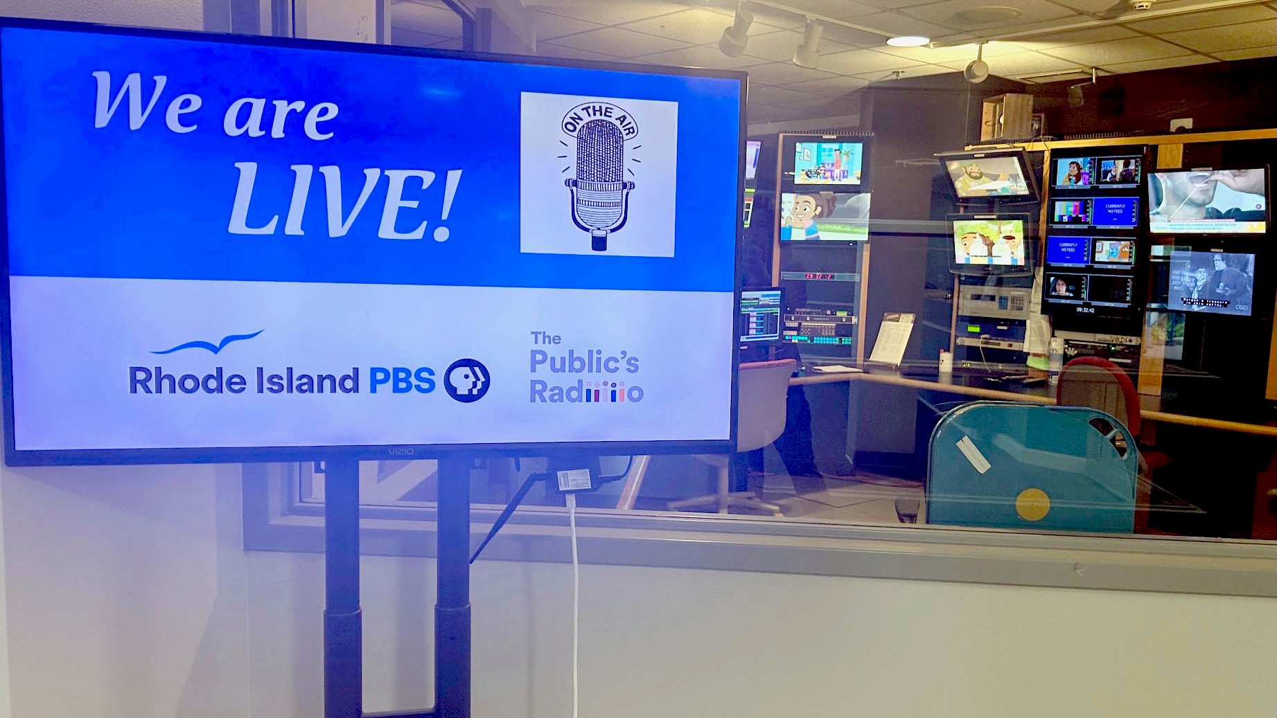 A television monitor in a control room displays the message “We are LIVE!” with a vintage microphone graphic and the words “On the Air.” Logos for Rhode Island PBS and The Public’s Radio appear below. Behind the monitor, through a window, television monitors and broadcasting equipment fill the control room.