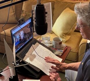 Scott Carter, host of Ye Gods, sits at a desk with headphones on, speaking into a microphone while recording an interview with Martin Short via video call on his laptop. Papers, a mug, and a glass of water are on the table, and a ring light and microphone are set up for the recording.