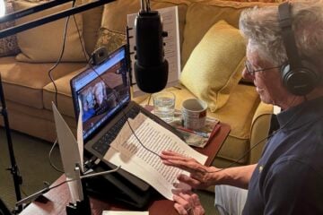 Scott Carter, host of Ye Gods, sits at a desk with headphones on, speaking into a microphone while recording an interview with Martin Short via video call on his laptop. Papers, a mug, and a glass of water are on the table, and a ring light and microphone are set up for the recording.