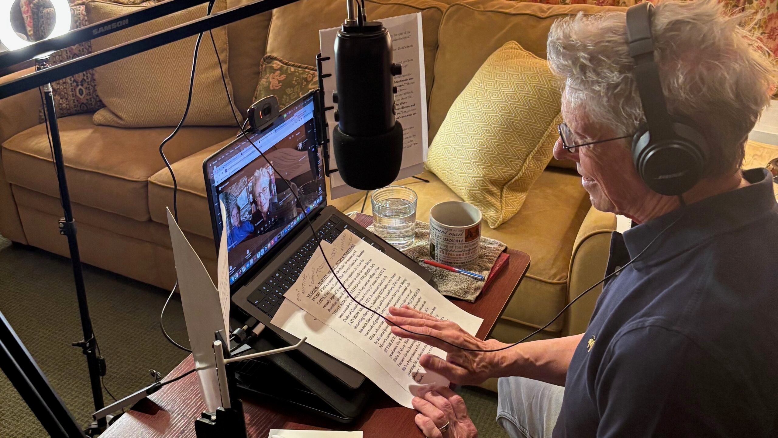Scott Carter, host of Ye Gods, sits at a desk with headphones on, speaking into a microphone while recording an interview with Martin Short via video call on his laptop. Papers, a mug, and a glass of water are on the table, and a ring light and microphone are set up for the recording.