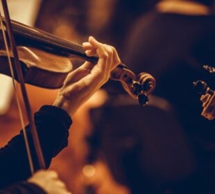 A close-up of a musician’s hand playing a violin, fingers pressing the strings on the fingerboard while the bow moves across the strings. The warm lighting highlights the wood grain of the instrument and creates a soft, intimate atmosphere.