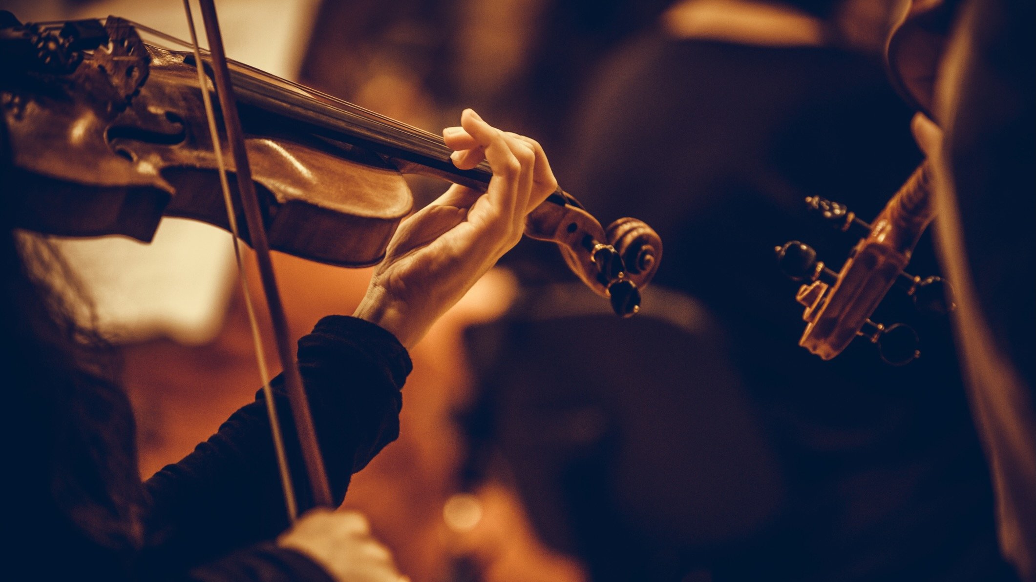 A close-up of a musician’s hand playing a violin, fingers pressing the strings on the fingerboard while the bow moves across the strings. The warm lighting highlights the wood grain of the instrument and creates a soft, intimate atmosphere.
