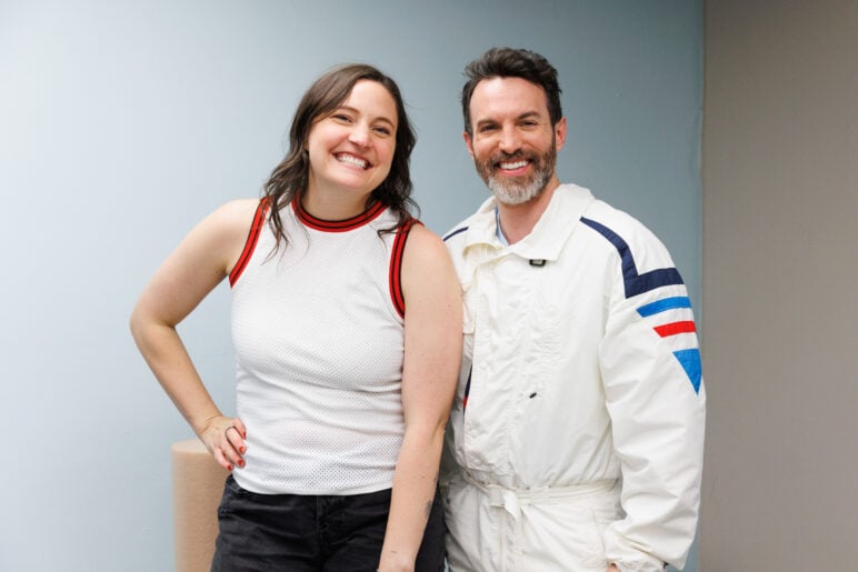 Erin Newins and Tyler Coe smile while posing together indoors against a light blue wall. Newins wears a sleeveless white top with red trim, and Coe wears a white jacket with blue and red stripes on the sleeves.