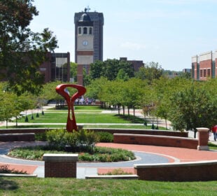 A landscaped central walkway on Western Kentucky University’s campus, with green lawns, trees, and red-brick paths. A modern red abstract sculpture sits in the foreground within a circular garden. In the background, the Guthrie Bell Tower with its clock face rises above campus buildings, while groups of people walk along the tree-lined paths.