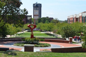 A landscaped central walkway on Western Kentucky University’s campus, with green lawns, trees, and red-brick paths. A modern red abstract sculpture sits in the foreground within a circular garden. In the background, the Guthrie Bell Tower with its clock face rises above campus buildings, while groups of people walk along the tree-lined paths.