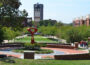 A landscaped central walkway on Western Kentucky University’s campus, with green lawns, trees, and red-brick paths. A modern red abstract sculpture sits in the foreground within a circular garden. In the background, the Guthrie Bell Tower with its clock face rises above campus buildings, while groups of people walk along the tree-lined paths.