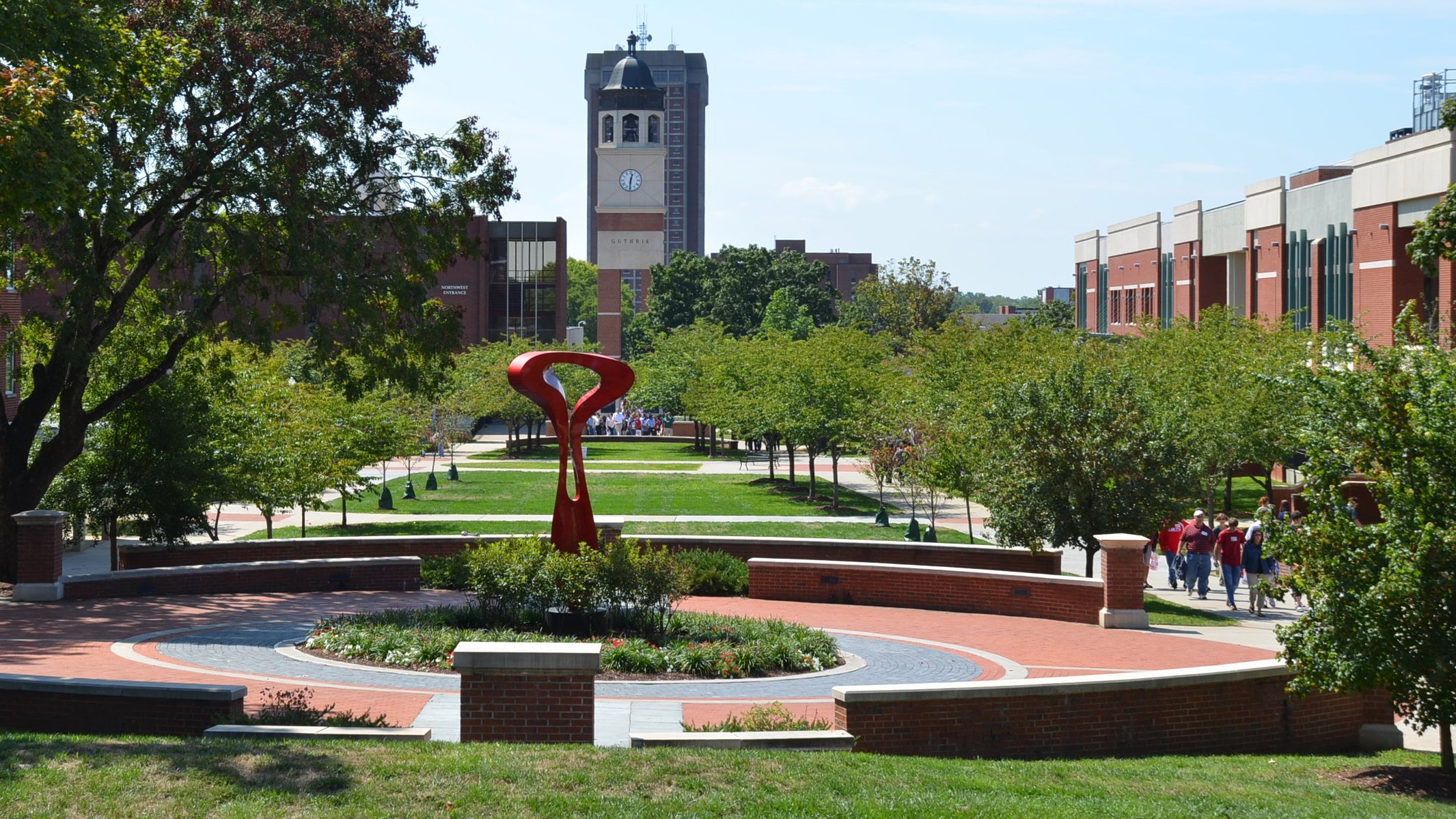 A landscaped central walkway on Western Kentucky University’s campus, with green lawns, trees, and red-brick paths. A modern red abstract sculpture sits in the foreground within a circular garden. In the background, the Guthrie Bell Tower with its clock face rises above campus buildings, while groups of people walk along the tree-lined paths.