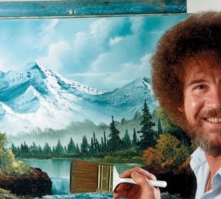 Bob Ross smiling beside a canvas depicting snowcapped mountains, pine trees, and a lake, holding a large brush in front of his landscape painting.