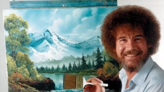 Bob Ross smiling beside a canvas depicting snowcapped mountains, pine trees, and a lake, holding a large brush in front of his landscape painting.