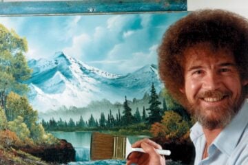 Bob Ross smiling beside a canvas depicting snowcapped mountains, pine trees, and a lake, holding a large brush in front of his landscape painting.