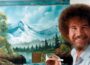 Bob Ross smiling beside a canvas depicting snowcapped mountains, pine trees, and a lake, holding a large brush in front of his landscape painting.