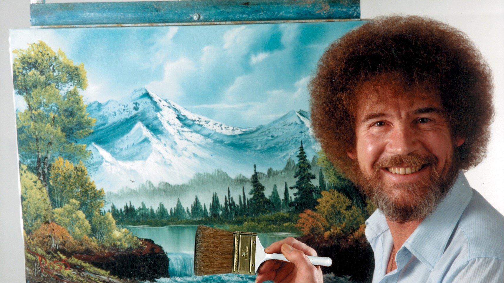 Bob Ross smiling beside a canvas depicting snowcapped mountains, pine trees, and a lake, holding a large brush in front of his landscape painting.