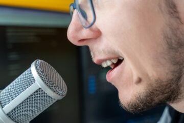 Close-up of a person speaking into a studio microphone, showing their mouth and part of their face in front of a blurred background.