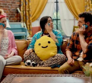 On an episode of How Are We Today?, Tyler, right, sits on a couch with his neighbors Barbara and Mariel. All three hold blue ice packs to their faces as part of an exercise for managing emotions. The scene takes place in a cozy, brightly colored living room with a brick wall, yellow curtains, and a blue piano in the background.