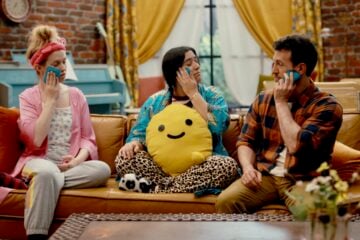 On an episode of How Are We Today?, Tyler, right, sits on a couch with his neighbors Barbara and Mariel. All three hold blue ice packs to their faces as part of an exercise for managing emotions. The scene takes place in a cozy, brightly colored living room with a brick wall, yellow curtains, and a blue piano in the background.