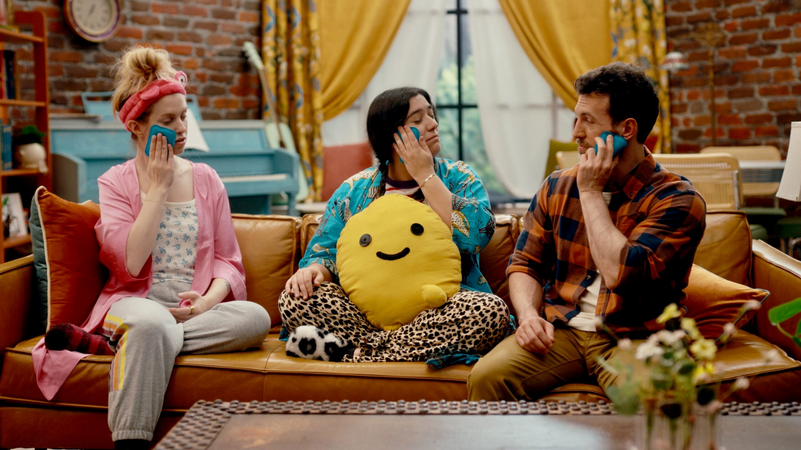 On an episode of How Are We Today?, Tyler, right, sits on a couch with his neighbors Barbara and Mariel. All three hold blue ice packs to their faces as part of an exercise for managing emotions. The scene takes place in a cozy, brightly colored living room with a brick wall, yellow curtains, and a blue piano in the background.