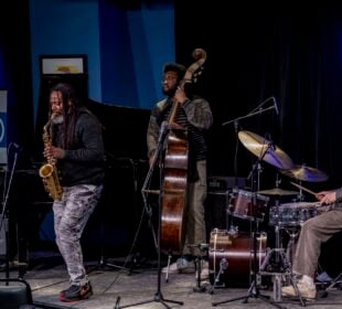 A jazz quartet performs on stage at the Bop Stop in Cleveland. A saxophonist stands at center playing beside a pianist, an upright bassist, and a drummer. Audience members watch from the foreground as the musicians play under blue lighting and a neon Bop Stop sign.