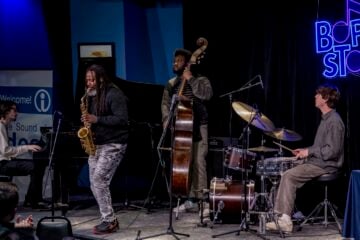 A jazz quartet performs on stage at the Bop Stop in Cleveland. A saxophonist stands at center playing beside a pianist, an upright bassist, and a drummer. Audience members watch from the foreground as the musicians play under blue lighting and a neon Bop Stop sign.
