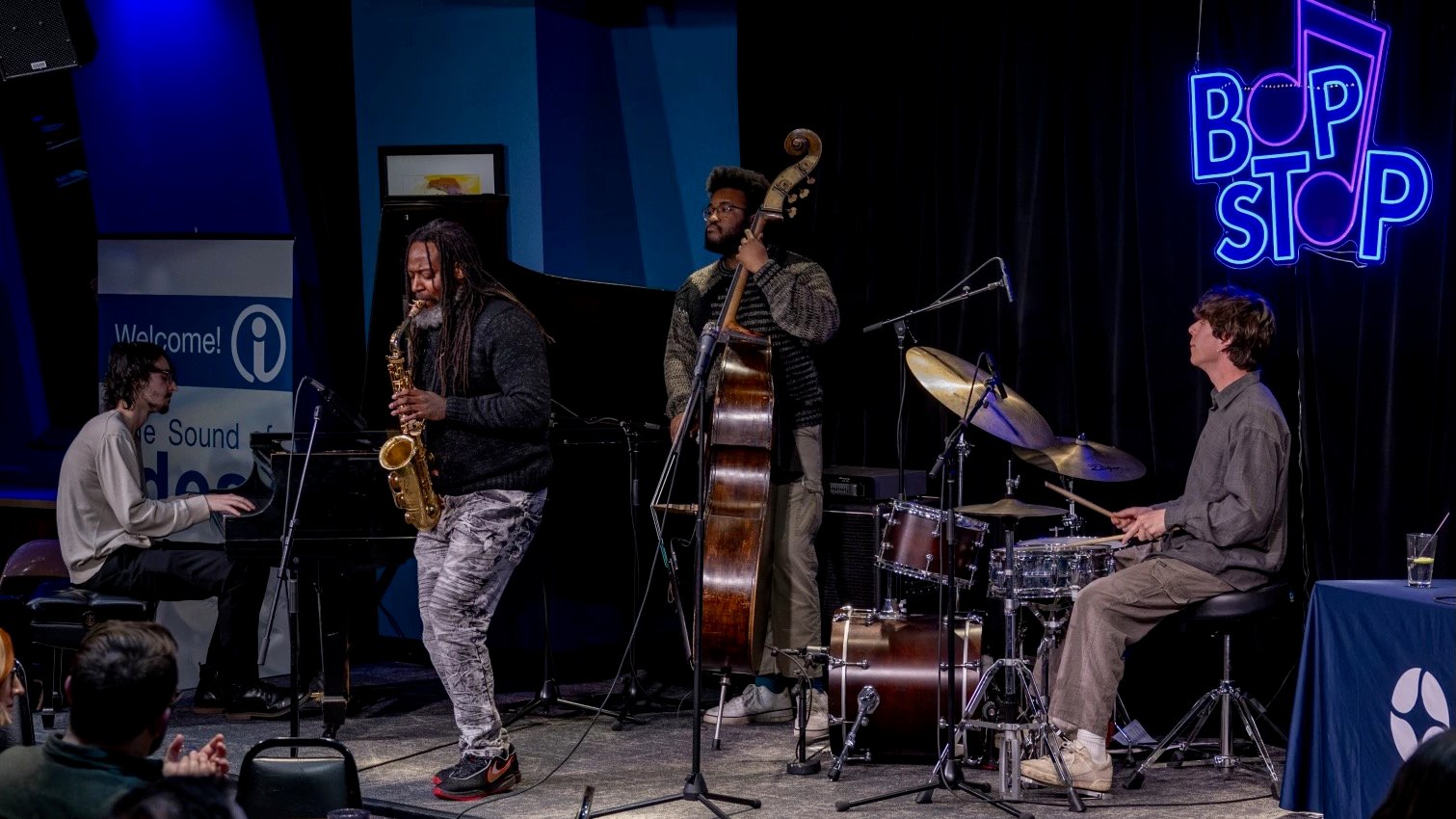 A jazz quartet performs on stage at the Bop Stop in Cleveland. A saxophonist stands at center playing beside a pianist, an upright bassist, and a drummer. Audience members watch from the foreground as the musicians play under blue lighting and a neon Bop Stop sign.