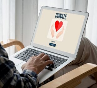 A person seated indoors uses a laptop displaying a donation webpage with an image of hands holding a red heart and a button that says “Support Us.”