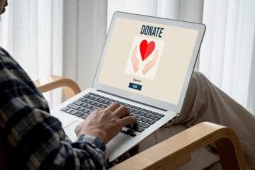 A person seated indoors uses a laptop displaying a donation webpage with an image of hands holding a red heart and a button that says “Support Us.”