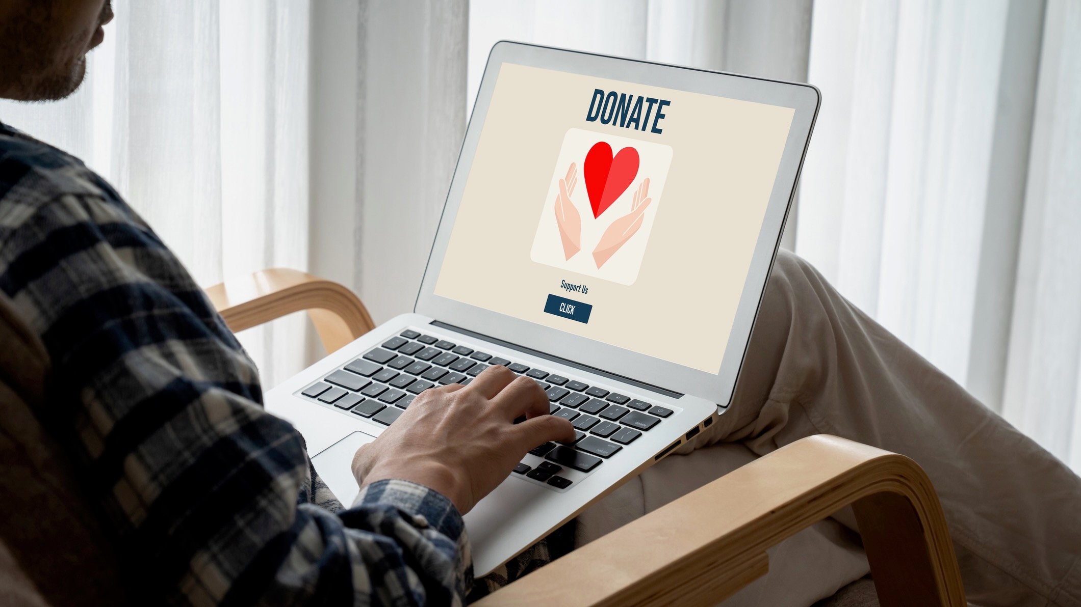 A person seated indoors uses a laptop displaying a donation webpage with an image of hands holding a red heart and a button that says “Support Us.”