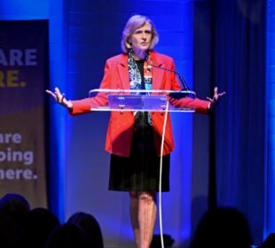 PBS President and CEO Paula Kerger stands behind a clear lectern with arms outstretched while speaking about the future of public media at the Sanctuary Events Center in Fargo. Blue stage lighting and a banner reading “We are here. We are not going anywhere.” frame the Oct. 2, 2025, event.
