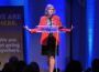 PBS President and CEO Paula Kerger stands behind a clear lectern with arms outstretched while speaking about the future of public media at the Sanctuary Events Center in Fargo. Blue stage lighting and a banner reading “We are here. We are not going anywhere.” frame the Oct. 2, 2025, event.