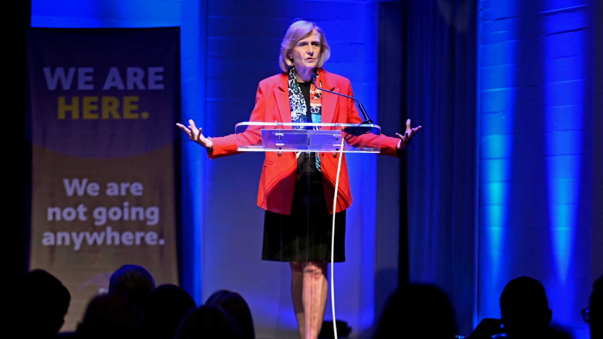 PBS President and CEO Paula Kerger stands behind a clear lectern with arms outstretched while speaking about the future of public media at the Sanctuary Events Center in Fargo. Blue stage lighting and a banner reading “We are here. We are not going anywhere.” frame the Oct. 2, 2025, event.