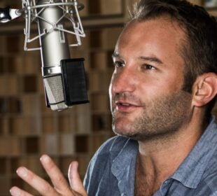 David Greene speaks into a large studio microphone, gesturing with one hand as he talks. He is seated in a recording booth with a blurred background of wooden acoustic panels.
