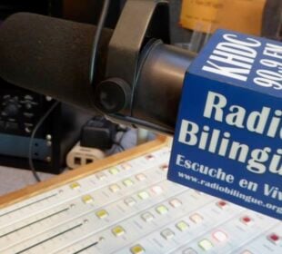 A close-up of a studio microphone with a blue mic flag labeled “Radio Bilingüe KHDC 90.9 FM, Salinas, CA,” positioned above a broadcast mixing console with lit buttons and audio equipment in the background.